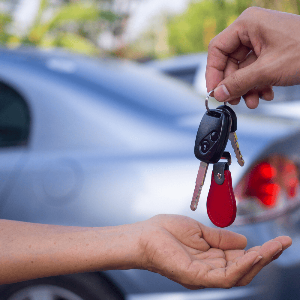 Set of car keys being passed from one hand to another with a silver car in the background.