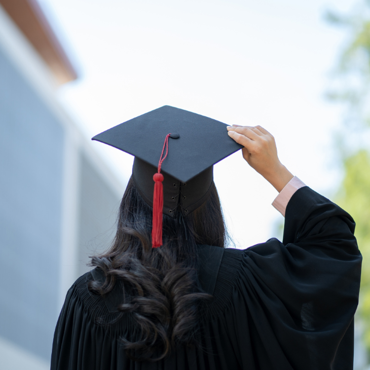 woman from behind with dark hair wearing graduation cap and gown with one hand on cap