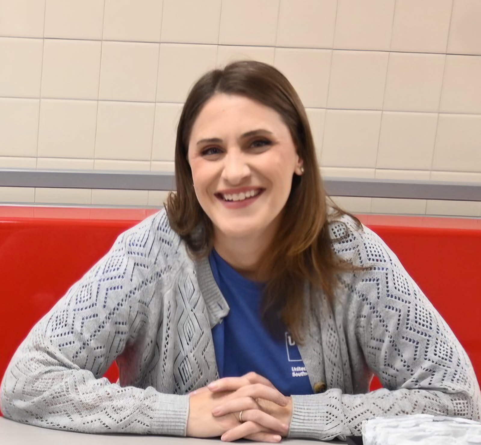 Woman with brown hair wearing a grey sweater ad blue shirt smiling with hands in front of her while sitting in a red booth.
