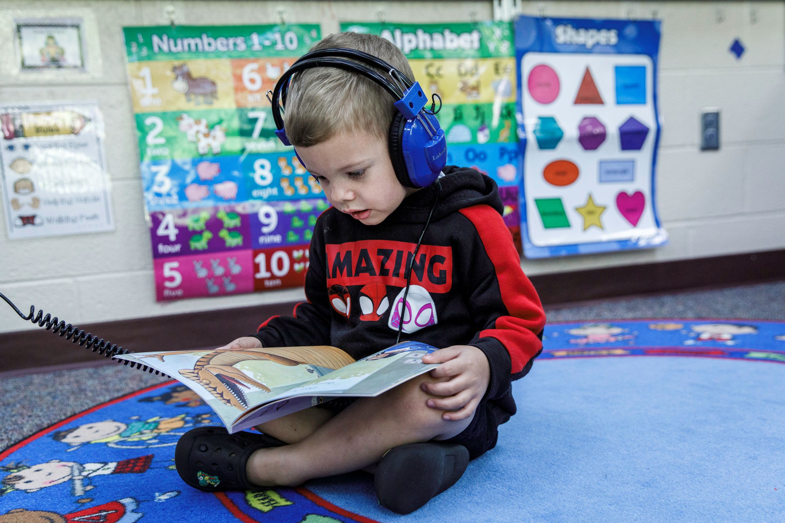 A young boy reading a book with blue headphones on.